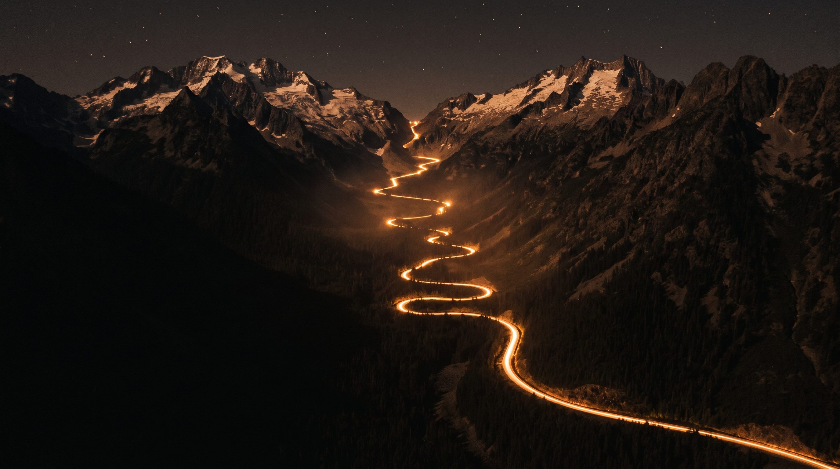 Night aerial of a mountain pass. Snow-capped peaks hold a faint warm afterglow under the stars, and one amber light trail climbs the switchbacks up the valley.